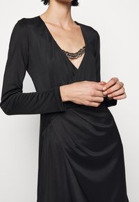 Schwarzes Kleid aus glattem Stoff, mit einem tiefen V-Ausschnitt, der mit einem Kettendetail akzentuiert ist, und langen Ärmeln. Der Körper hat eine drapierte Textur.
