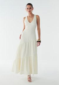 Lange, gelaagde jurk in crème, met een V-hals, een aangesloten lijfje, gestructureerde stof en een vloeiende rok. Draag gecombineerd met sandalen met bandjes en armbanden.