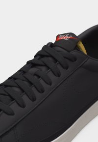 Baskets basses noires avec lacets noirs, un discret logo Swoosh cousu, semelle blanche, logo rouge sur la languette et doublure intérieure jaune.