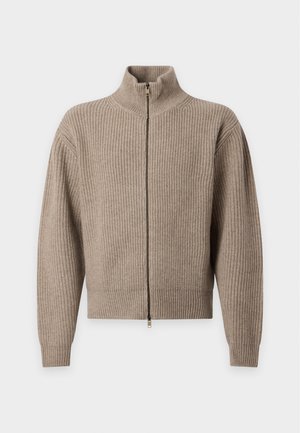 Ung mand iført en beige strikkejakke med lynlås, hvid skjorte, løse sorte bukser og hvide sneakers står mod en ensfarvet lys baggrund.