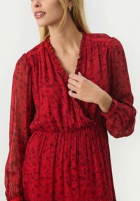 Rode jurk met bloemenpatroon, voorzien van een rimpelachtige halslijn, elastische taille en doorschijnende lange mouwen; ingewikkelde zwarte details op de stof.