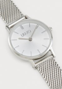 Orologio argento con quadrante rotondo in metallo spazzolato, indicatori delle ore minimalisti e cinturino in acciaio inossidabile a maglia. Design durevole ed elegante.