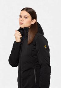 Giacca imbottita nera con cappuccio, caratterizzata da un colletto alto, tasche laterali con zip e una toppa con logo sulla spalla sinistra. Texture liscia.