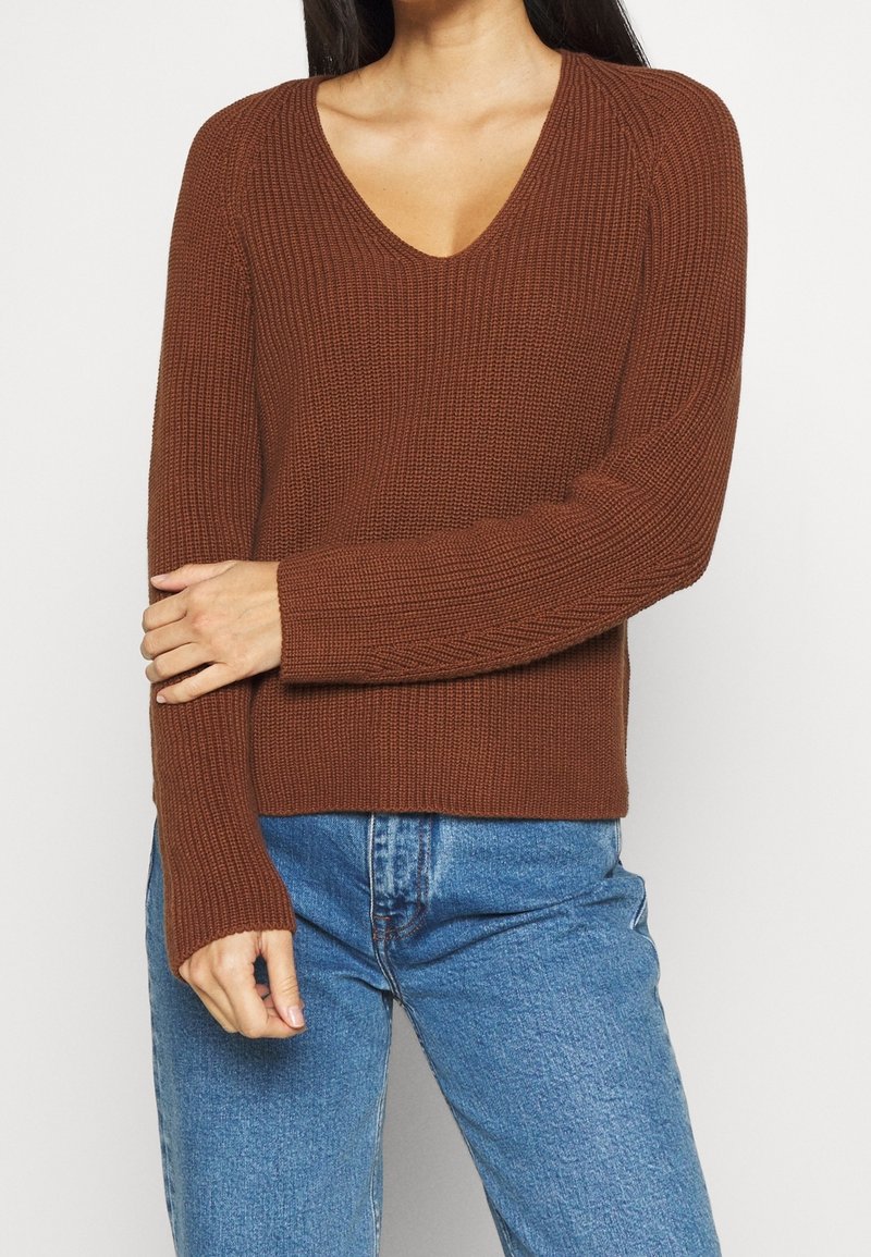 Hnědý pletený svetr s výstřihem do V, ribbovanou strukturou a dlouhými rukávy. Kombinovaný s světle modrými džíny, což vytváří ležérní vzhled.