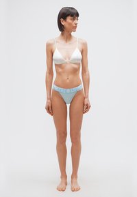 Γυναίκα όρθια ξυπόλυτη, φοράει λευκό τρίγωνο σουτιέν και ανοιχτό μπλε μπικίνι με σχέδια, κοιτάζοντας προς τα αριστερά σε λευκό φόντο.