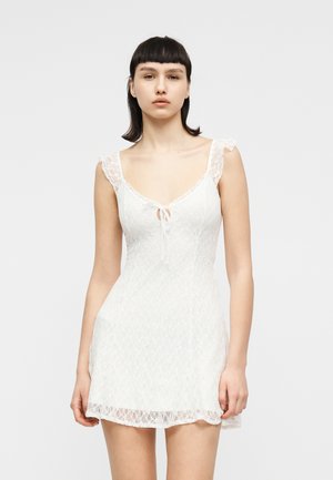 Kobieta ubrana w krótką białą koronkową sukienkę bez rękawów z małą wiązaną kokardą z przodu, stojąca na tle jednolitego jasnoszarego tła.