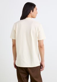 T-shirt a maniche corte color crema con scollatura rotonda e spalle senza cuciture, abbinato a pantaloni marroni. Texture liscia.