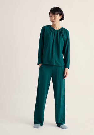 Femme aux cheveux courts noirs portant un haut à manches longues boutonné vert sarcelle foncé et un pantalon ample assorti, debout les mains dans les poches sur un fond clair.