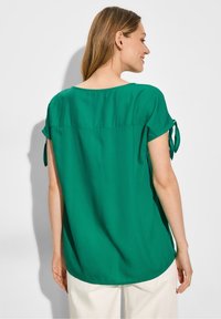 Blusa verde a maniche corte con orlo arrotondato e fiocchi laterali. Realizzata in tessuto leggero con una superficie liscia. Vista posteriore mostrata.