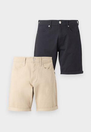 Młody mężczyzna stoi, ubrany w białą koszulkę Pepe Jeans, czarne szorty, białe skarpetki i czarne trampki na jednolitym białym tle.