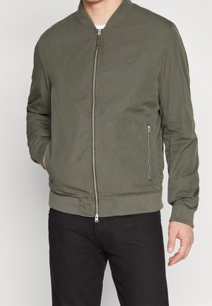 Homme portant une veste bomber vert olive avec fermeture éclair à l'avant et poches latérales, associée à un pantalon noir, debout devant un fond uni.