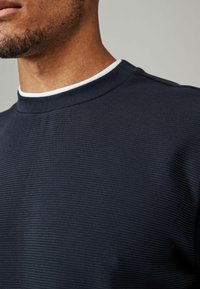 Granatowy sweatshirt z wyraźnym prążkowanym wzorem, okrągłym dekoltem z białym akcentem i wytrzymałym materiałem, nadający nowoczesny wygląd.