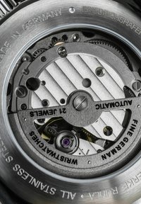 Automatic watch movement with silver-toned metal components, engraved text, circular gears, and a purple jewel accent. Features intricate textures.