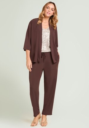 Frau trägt braune, locker sitzende Jacke und Hose mit silbernem Pailletten-Top, stehend vor einem einfarbigen hellen Hintergrund.