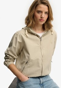 Chaqueta ligera beige con capucha, con puños elásticos, bolsillos laterales y cremallera de longitud completa. Diseño minimalista con un logo sutil.