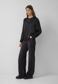 Kobieta stoi na jednolitym tle, ubrana w czarną, wzorzystą bluzkę z długim rękawem, szerokie ciemne spodnie i czarne buty.