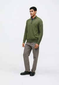 Πράσινο πλεκτό πουλόβερ με γιακά μισού φερμουάρ, συνδυασμένο με γκρι παντελόνι και μαύρα παπούτσια. Το μοντέλο στέκεται σε ουδέτερο φόντο, κοιτάζοντας ελαφρώς προς το πλάι.