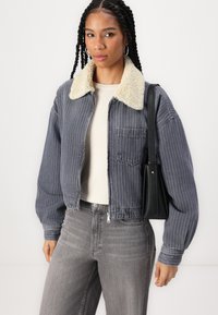 Gråstripet denimjakke med krage i kremfarget kunstpels, lommedetaljer og elastisk nederkant. Kombinert med en lys topp og grå jeans.