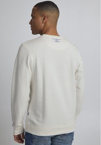 Sudadera de algodón blanca con mangas largas, puños de canalé y dobladillo. Presenta una etiqueta en la parte posterior del cuello con texto azul. Combinada con jeans azules.