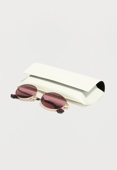 Στρογγυλά γυαλιά ηλίου με χρυσά πλαίσια και μοβ φακούς που ακουμπούν μπροστά από μια λευκή ορθογώνια θήκη γυαλιών πάνω σε μια απλή επιφάνεια.