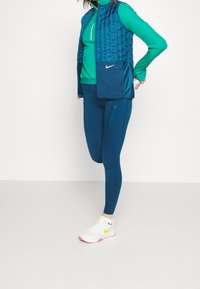 Osoba v teal dlouhém rukávu, námořnickém prošívaném vestě a námořnických legínách, natahující jednu nohu dozadu, v bílých teniskách Nike.