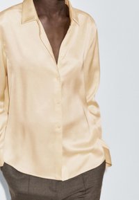 Chemise en satin beige clair avec une fermeture boutonnée à l'avant, manches longues et coupe décontractée. Texture lisse et léger éclat. À assortir avec un pantalon foncé.