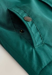Tissu vert, lisse, avec une poche à rabat, dotée d'une fermeture à bouton-pression en laiton. Des coutures soignées le long des bords renforcent la durabilité.