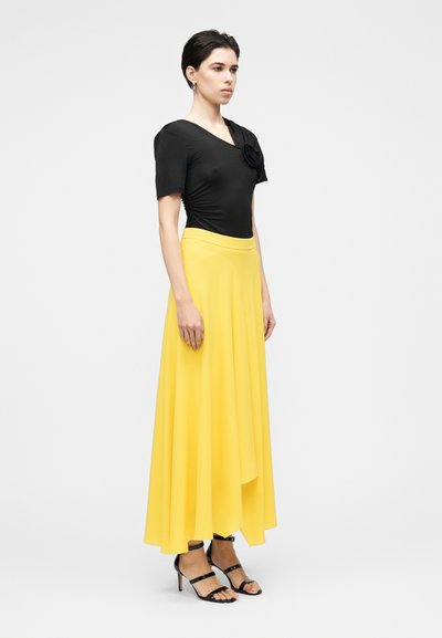 Haut noir à manches courtes avec un accent floral texturé, associé à une jupe maxi jaune fluide et des talons noirs à lanières. Tissu lisse, drapé élégant.