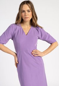 Lila Kleid mit kurzen Puffärmeln, V-Ausschnitt und taillierter Passform. Glatte Textur mit subtilen Nahtdetails. Vordertaschen sichtbar.