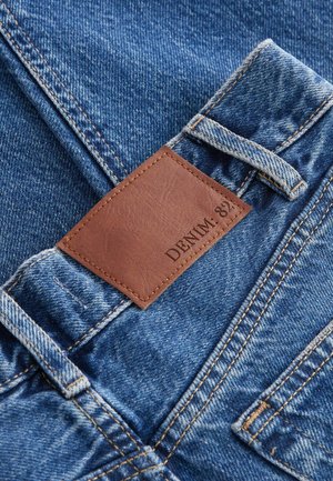 Jean en denim avec un tissu délavé bleu, présentant une étiquette en cuir marron avec l'inscription "DENIM : 821" et des détails de couture visibles.