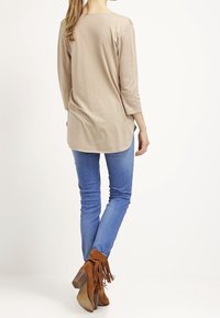 Top beige a maniche lunghe con orlo arrotondato, abbinato a jeans skinny azzurri e stivaletti marroni con frange, visto da dietro.