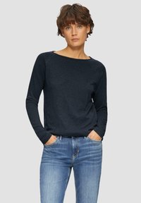 Frau mit kurzen braunen Haaren, die ein dunkelblaues Langarmshirt und eine bluejeans trägt, steht mit den Händen in den Taschen vor einem einfarbigen Hintergrund.