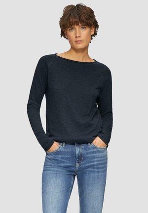 Femme avec les cheveux bruns courts portant une chemise à manches longues bleu marine et un jean bleu, se tenant avec les mains dans les poches devant un fond uni.