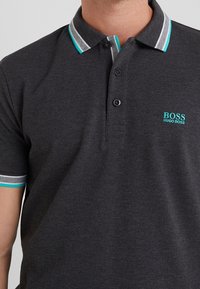 Mörkgrå pikétröja med tvåknappskrage, turkosa och randiga detaljer på kragen och ärmarna, samt en logotyp fram.