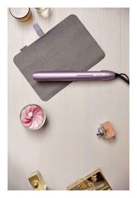 Lila Haarglätter auf einem grauen Filzmat. In der Nähe befinden sich ein Glas Getränk, eine Parfümflasche und eine dekorative Glasblume mit pinken Blütenblättern.