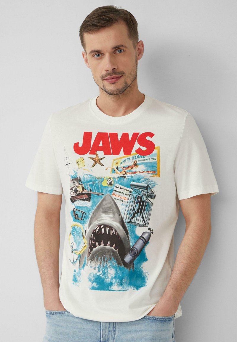 T-shirt en coton blanc présentant "JAWS" en lettres rouges audacieuses, avec un graphique de requin, des vagues océaniques et des détails liés à la plage. Coupe décontractée.