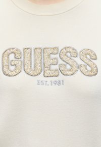 Tissu couleur crème avec « GUESS » écrit en grandes lettres ornées et « EST. 1981 » brodé en dessous.