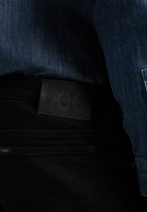 Veste en denim rentrée dans un jean noir Lee montrant l'écusson de la marque Lee et l'étiquette sur la ceinture arrière et la poche.