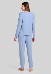 Mujer de pie descalza, de espaldas, vistiendo una blusa de manga larga azul claro y pantalones a juego, contra un fondo gris uniforme.