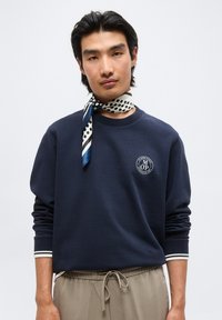 Άνδρας που φοράει μπλε σκούρο φούτερ με λογότυπο, μπεζ παντελόνι με κορδόνι και λευκό κασκόλ με πουά και ρίγες δεμένο γύρω από το λαιμό του.