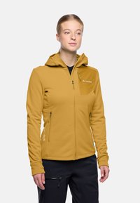 Giacca softshell gialla con cappuccio, dotata di tasche con zip e tasca sul petto. Le cuciture piatte e la texture suggeriscono flessibilità e comfort.