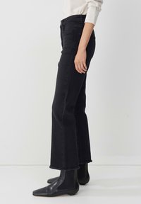 Black flared jeans with a high waist, made of denim. Features a raw hem and side pockets, paired with sleek black ankle boots.