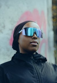 Γυαλιά ηλίου με μεγάλο, γωνιώδες σκελετό σε λαμπερό ασημί και φακούς με αντανακλαστικό βαθμιαίο χρωματισμό. Φοριούνται με μαύρο μπουφάν και καλυμμα κεφαλής.