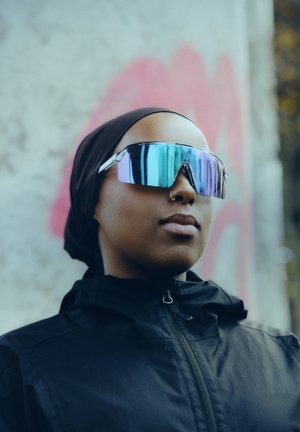 Slnečné okuliare s nadrozmerným, uhlovým rámom v lesklej striebornej farbe a reflektujúcimi šošovkami s prechodom. Nosia sa s čiernou bundou a pokrývkou hlavy.