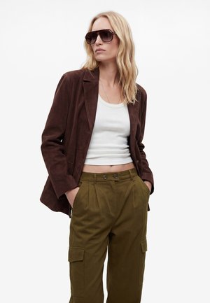 Frau mit blondem Haar, die dunkle Sonnenbrillen, einen braunen Blazer, ein weißes kurzes Top und olivfarbene Cargohosen trägt, mit den Händen in den Taschen.