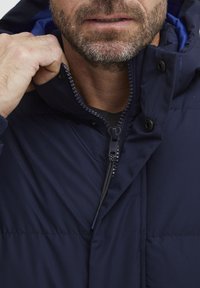 Marineblå jakke med fylling, med glidelås, trykknapper og høy krage. Teksturert stoff og sømdetaljer er synlige.