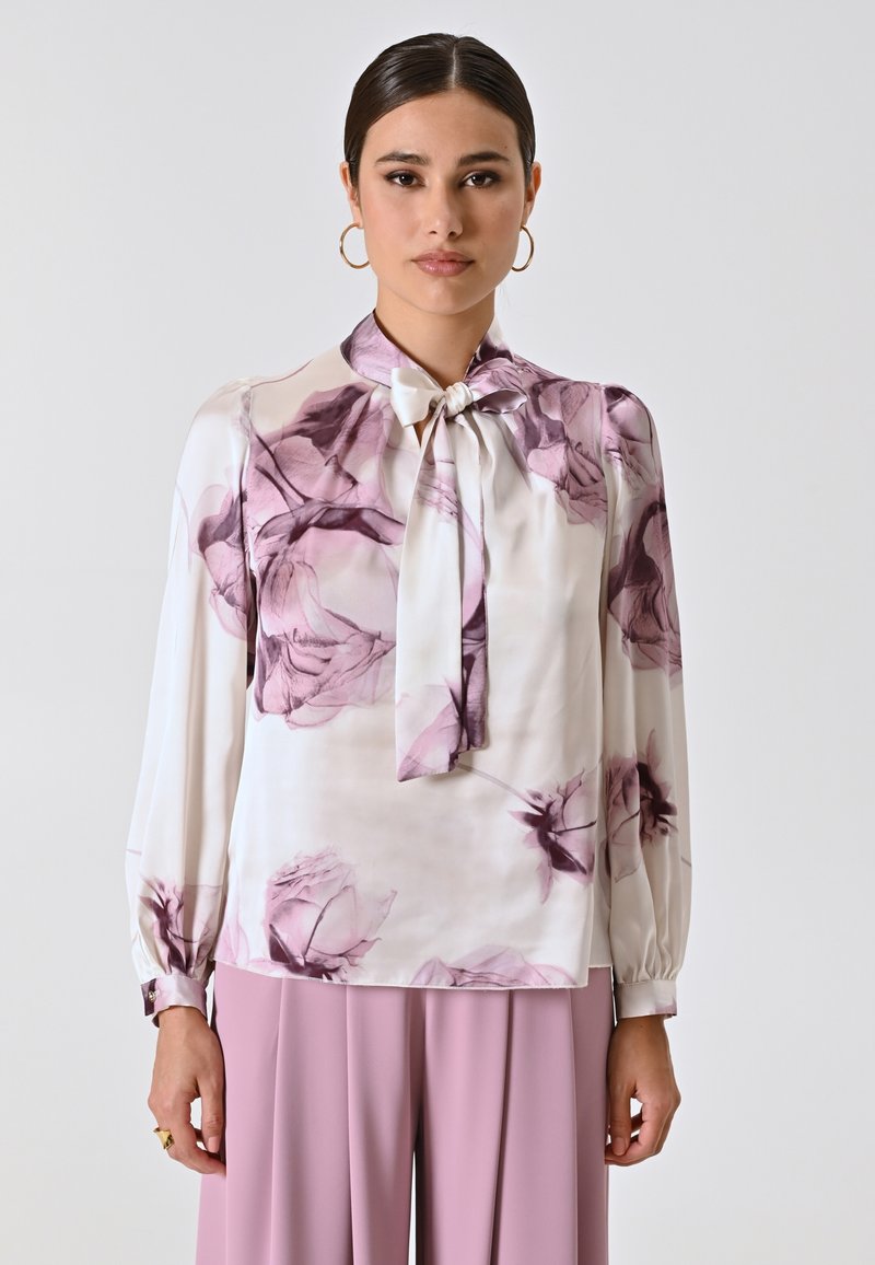 Blusa bianca a maniche lunghe con stampa floreale viola e colletto a fiocco. Tessuto morbido con una finitura liscia, abbinata a pantaloni rosa chiaro.