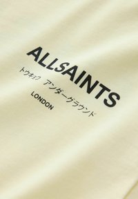 Text ALLSAINTS na krémovej látke s japonskými znakmi a slovom LONDON vytlačeným pod ním čiernou farbou.