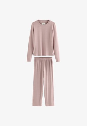 Mujer con un top de manga larga color mauve y pantalones de pierna ancha apoyada en la encimera de la cocina, con el fregadero, estantes, tazas y cuencos visibles en el fondo.