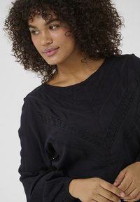 Black blouse featuring a wide neckline, textured accents in a chevron pattern, and long sleeves with gathered cuffs. Smooth fabric finish.